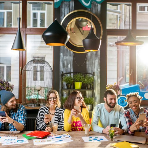 Multi ethnic group of young friends sitting with phones at the big table in the modern cafe interior